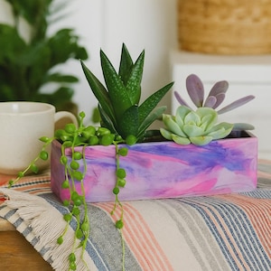 Puede incluir: Una jardinera rectangular con un diseño veteado rosa y morado, llena de varias suculentas y plantas colgantes. La jardinera se encuentra sobre una tela a rayas, mostrando una estética decorativa moderna.
