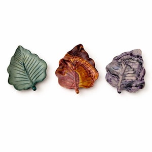 May include: Three leaf-shaped dishes arranged in a row. The first dish is a matte sage green, the second features a marbled blend of deep amber and brown tones, and the third displays a swirling pattern of dark purple and light gray hues.