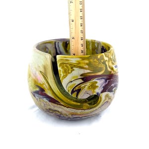 May include: A handcrafted yarn bowl with a swirling, marbled design in shades of green, gold, and purple. The bowl is approximately 4 inches tall, with a curved slot to hold yarn. The interior is smooth, and the exterior has a textured finish.