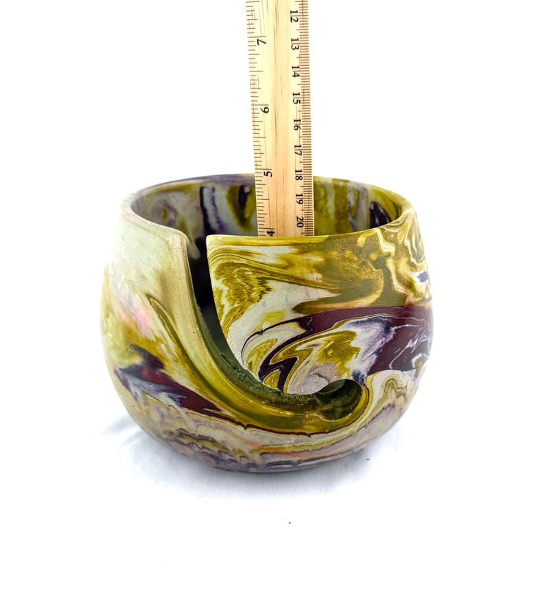 May include: A handcrafted yarn bowl with a swirling, marbled design in shades of green, gold, and purple. The bowl is approximately 4 inches tall, with a curved slot to hold yarn. The interior is smooth, and the exterior has a textured finish.