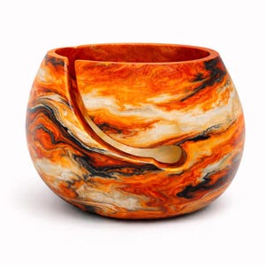 May include: A round yarn bowl with a swirling pattern of orange, black, and white. The bowl has a curved slot for the yarn to pass through. The bowl is a decorative item for knitters and crocheters.