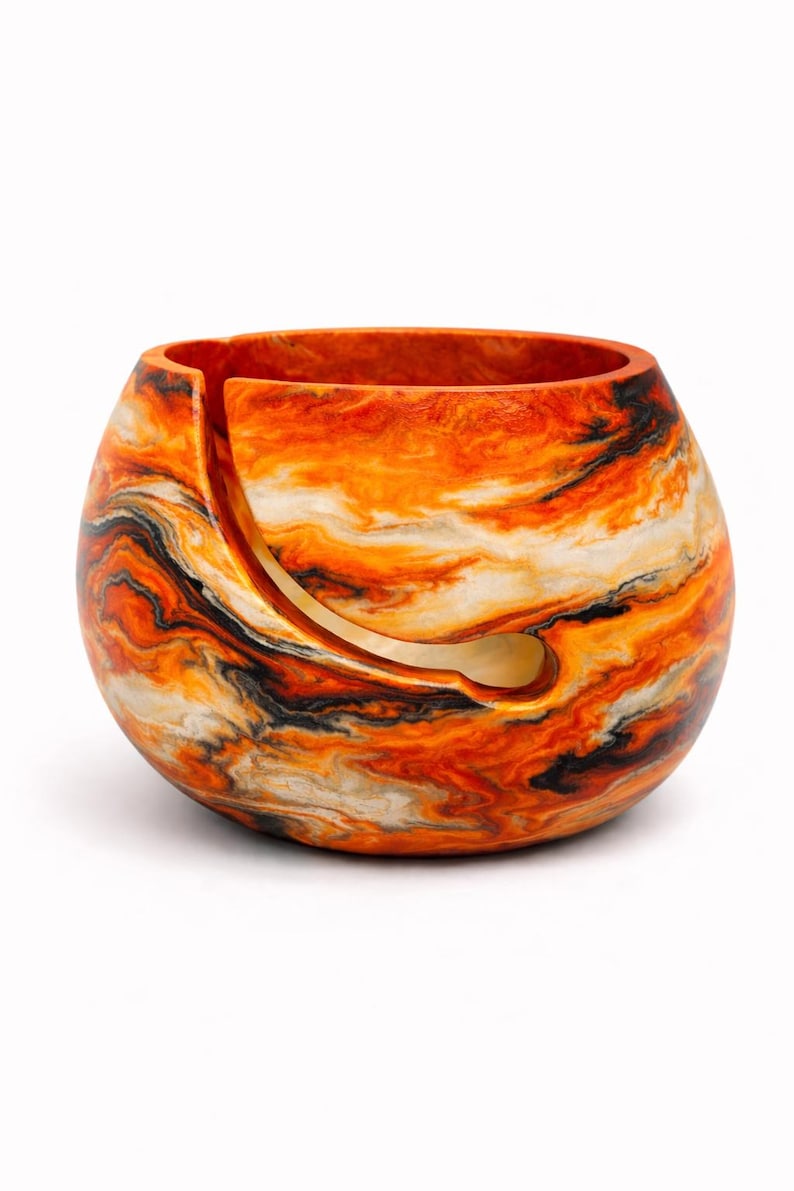 May include: A round yarn bowl with a swirling pattern of orange, black, and white. The bowl has a curved slot for the yarn to pass through. The bowl is a decorative item for knitters and crocheters.