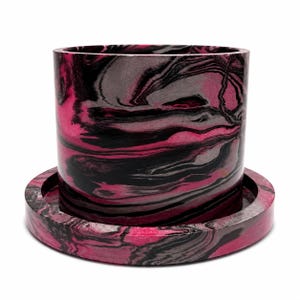 May include: A cylindrical planter with a matching circular saucer, both featuring a marbled pattern of vibrant hot pink, deep black, and soft grey swirls. The container has straight vertical walls and a smooth finish throughout the exterior.