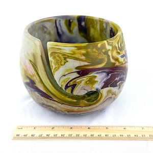 May include: A marbled yarn bowl in shades of olive green, purple, and cream. The bowl has a curved slot for yarn and a rounded shape. A wooden ruler is at the bottom of the image, measuring up to 12 inches.