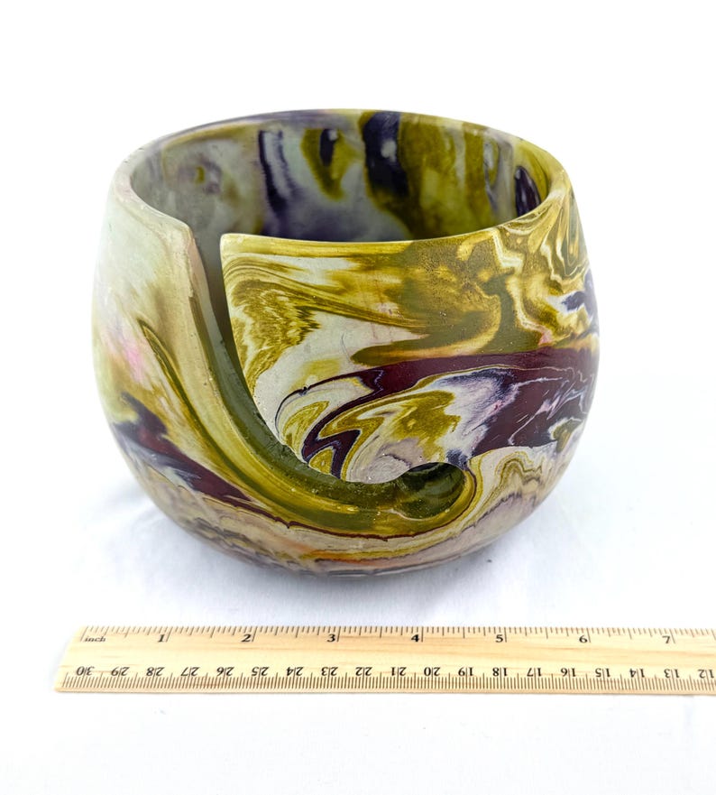 May include: A marbled yarn bowl in shades of olive green, purple, and cream. The bowl has a curved slot for yarn and a rounded shape. A wooden ruler is at the bottom of the image, measuring up to 12 inches.