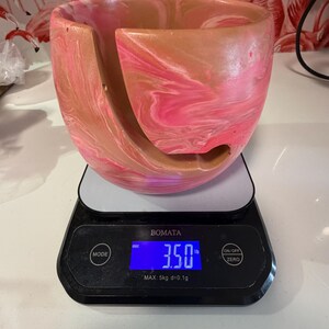 Handmade yarn bowl with soft pink and tan marbled swirl pattern, shown on a digital scale reading about 3.5 lb to demonstrate its ideal weight for knitting and crochet. Heavy enough to stay in place while yarn feeds smoothly, yet light enough to remain portable for crafting anywhere. This weighted yarn holder features a curved yarn guide slot to prevent tangles, making it perfect for knitters, crocheters, fiber artists, and thoughtful handmade gift shoppers.
