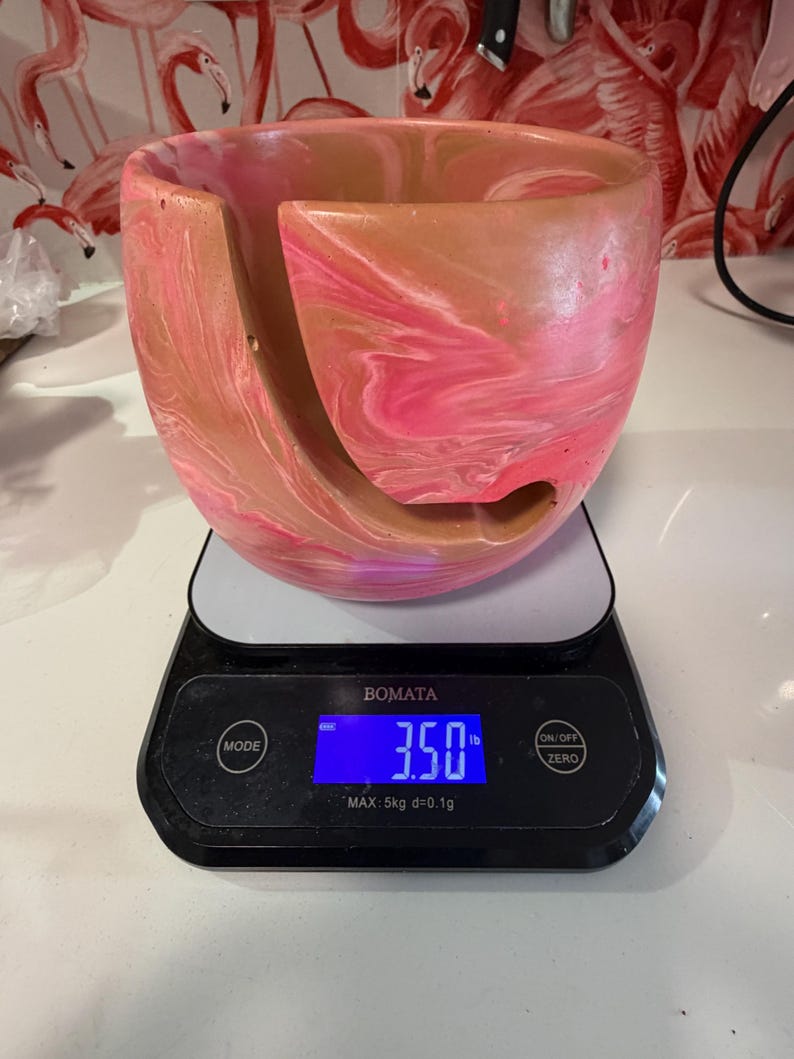 Handmade yarn bowl with soft pink and tan marbled swirl pattern, shown on a digital scale reading about 3.5 lb to demonstrate its ideal weight for knitting and crochet. Heavy enough to stay in place while yarn feeds smoothly, yet light enough to remain portable for crafting anywhere. This weighted yarn holder features a curved yarn guide slot to prevent tangles, making it perfect for knitters, crocheters, fiber artists, and thoughtful handmade gift shoppers.