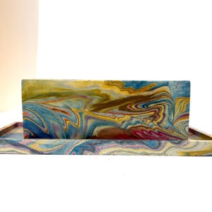 Puede incluir: Un juego decorativo de caja y bandeja con efecto marmolado. La caja es rectangular y vertical, y la bandeja también es rectangular. Los colores incluyen azul, amarillo, marrón y rosa, con un patrón arremolinado. El conjunto está sobre un fondo blanco.