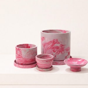 May include: A set of four decorative planters in varying sizes. The planters are cylindrical and bowl-shaped, with a pink and white marbled design. The set includes a small dish with a pedestal base.