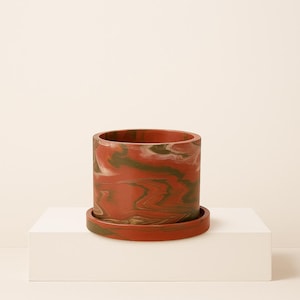 May include: A cylindrical planter with a matching saucer, featuring a marbled design in shades of red, green, and white. The planter is made of a solid material and sits on a white surface.
