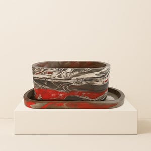Pode incluir: Um vaso retangular e uma bandeja a condizer com um design marmorizado. O vaso apresenta uma mistura de redemoinhos vermelhos, pretos e brancos. A bandeja é de cor e forma semelhantes, proporcionando um visual coeso para a decoração da casa.