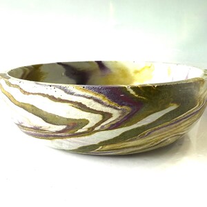 May include: A round bowl featuring a marbled pattern with swirls of olive green, deep purple, and off-white colors. The bowl has a smooth, curved exterior and a wide, open top, showcasing a continuous, fluid design across its entire surface.