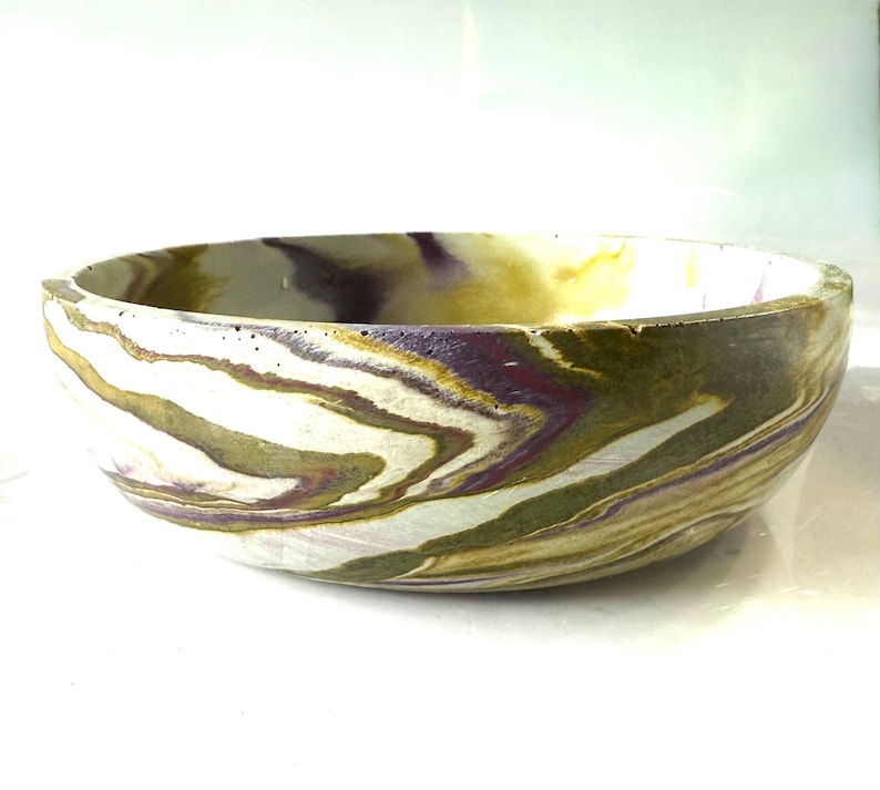 May include: A round bowl featuring a marbled pattern with swirls of olive green, deep purple, and off-white colors. The bowl has a smooth, curved exterior and a wide, open top, showcasing a continuous, fluid design across its entire surface.