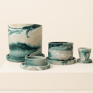 Puede incluir: Un conjunto de cinco macetas de cerámica de diferentes tamaños, cada una con un diseño único veteado en verde azulado y blanco. Las macetas incluyen platillos a juego. La maceta más grande mide unos 20 cm de alto.