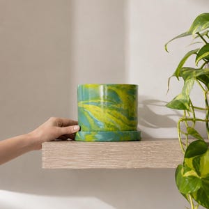 May include: A cylindrical ceramic plant pot with a glossy finish featuring a marbled pattern of vibrant lime green and deep teal swirls. The pot includes a matching circular saucer base, creating a cohesive look for indoor plant display.