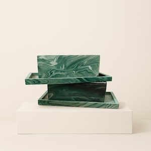 May include: Three stacked, rectangular trays with a marbled green and white pattern. The trays are different sizes and sit on a white rectangular base. The trays have a modern, minimalist design.