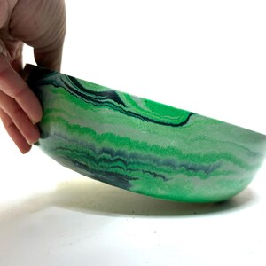 May include: A shallow, round bowl featuring a marbled pattern of vibrant green, light mint green, and dark charcoal gray swirls. The exterior surface displays a smooth, continuous flow of these colors, creating a layered, organic appearance across the curved sides of the vessel.