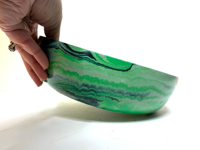 May include: A shallow, round bowl featuring a marbled pattern of vibrant green, light mint green, and dark charcoal gray swirls. The exterior surface displays a smooth, continuous flow of these colors, creating a layered, organic appearance across the curved sides of the vessel.