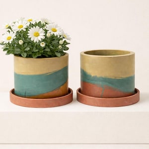 May include: Two cylindrical planters with a painted design. One contains a daisy plant. The planters are painted with bands of yellow, teal, and red-orange. Each planter sits on a matching terracotta-colored saucer.