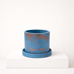 Puede incluir: Una maceta cilíndrica azul con un platillo a juego. La maceta tiene un efecto veteado de color cobre. El diseño es moderno y minimalista, adecuado para uso en interiores o exteriores. La maceta está hecha de un material duradero.