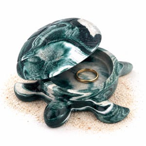 May include: A turtle-shaped trinket box crafted from dark green and white marbled stone. The shell lid is partially open, revealing a smooth interior basin that contains a single, plain gold-colored metal ring resting at the center of the base.