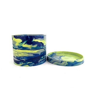 May include: A cylindrical container and a matching circular lid, both featuring a marbled pattern of deep blue, bright lime green, and grey swirls. The set has a smooth, solid finish with a consistent color blend across the exterior surfaces.