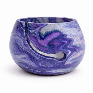 May include: A marbled yarn bowl in shades of purple, white, and blue. The bowl has a rounded shape with a curved slot for yarn to feed through. The swirling pattern gives it a unique, artistic look. A functional and decorative item for knitters and crocheters.