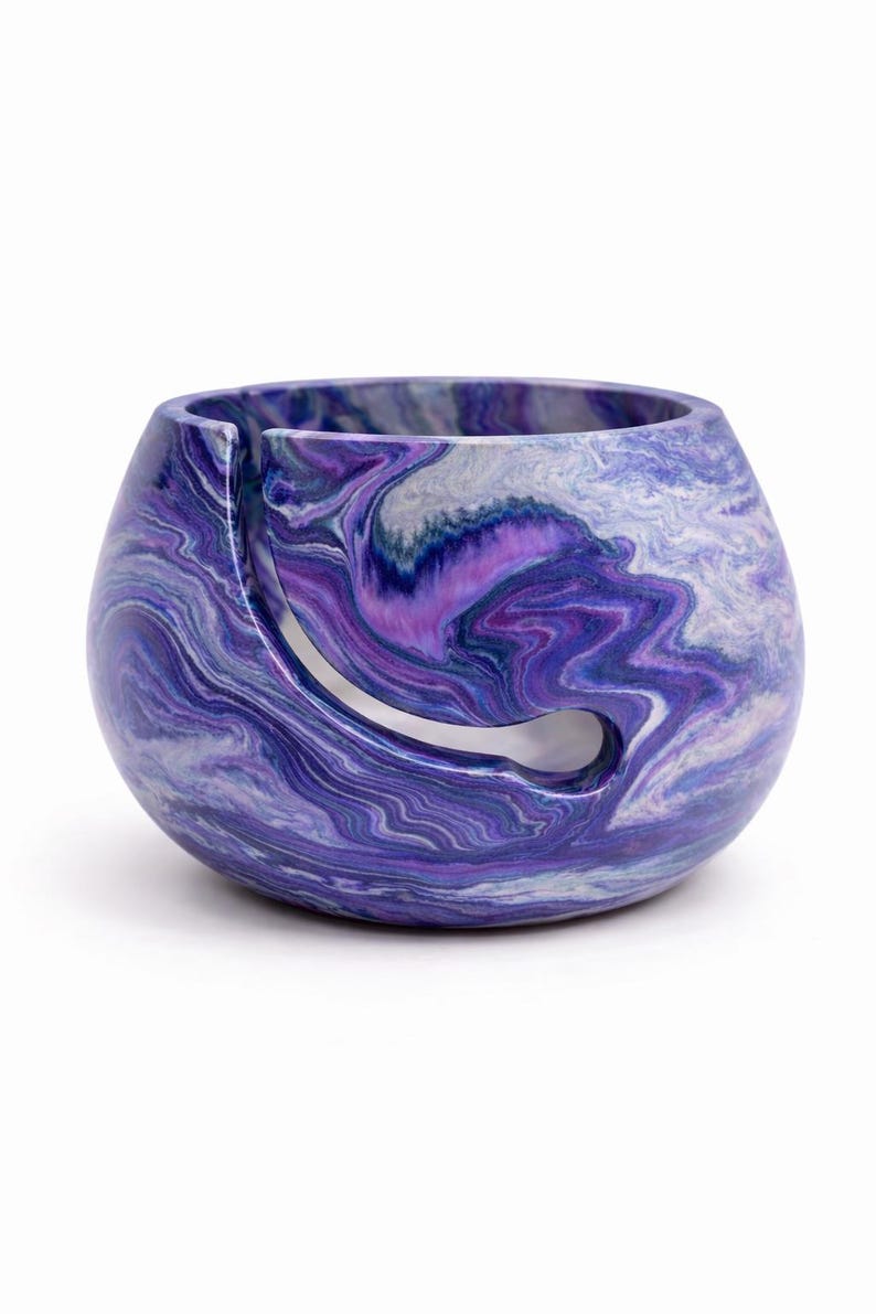 May include: A marbled yarn bowl in shades of purple, white, and blue. The bowl has a rounded shape with a curved slot for yarn to feed through. The swirling pattern gives it a unique, artistic look. A functional and decorative item for knitters and crocheters.