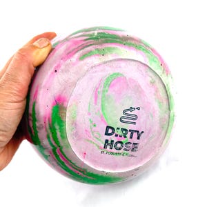 May include: A round, decorative object with a swirling pattern of pink, green, and white. The bottom features the text "DIRTY HOSE" and a hose graphic. The object is held by a hand against a white background.