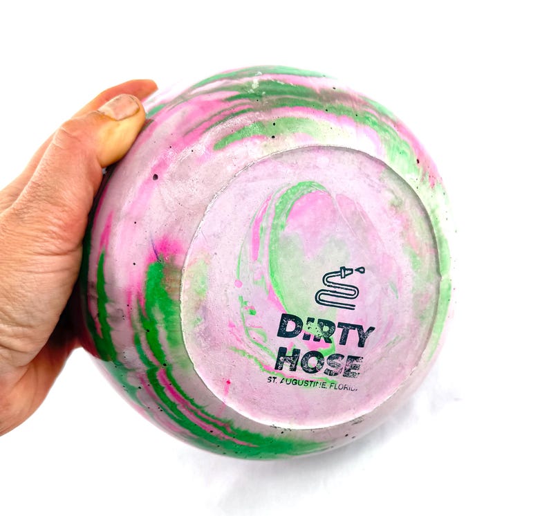 May include: A round, decorative object with a swirling pattern of pink, green, and white. The bottom features the text "DIRTY HOSE" and a hose graphic. The object is held by a hand against a white background.