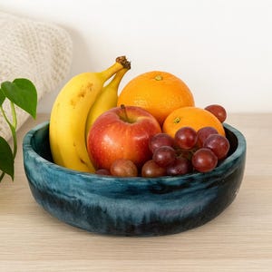 Op de afbeelding: Een blauwe keramische kom gevuld met vers fruit. De kom bevat bananen, een rode appel, sinaasappels en druiven. De kom staat op een licht houten oppervlak.
