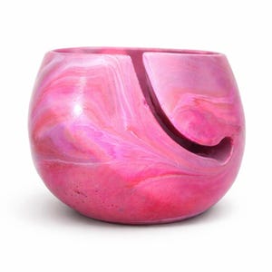 Handmade marbled yarn bowl in pink, coral, and soft white tones with a smooth glossy finish and curved yarn guide slot. This ceramic look yarn holder keeps yarn clean, untangled, and rolling smoothly while knitting or crocheting. Perfect for knitters, crocheters, fiber artists, cozy craft room decor. Unique swirl pattern makes each bowl one of a kind. Great gift for knitters, crochet gift for mom gift for crafters and handcrafted pottery collections.