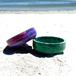 May include: Two decorative bowls on a sandy beach. One bowl is a vibrant mix of pink, purple, and blue. The other bowl is a solid green with a marbled effect. The bowls are round with a smooth finish.