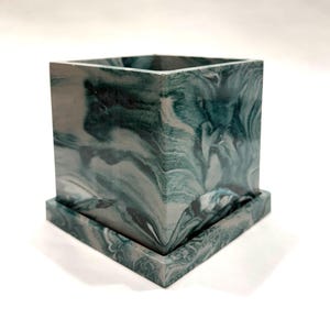 May include: A square planter with a matching square saucer, featuring a marbled pattern in shades of dark green, teal, and white. The container has straight vertical sides and a smooth finish, displaying a continuous swirling design across all surfaces.