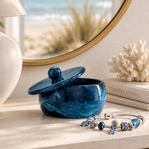 May include: A round, deep blue marbled trinket box with a matching lid and a silver-toned charm bracelet featuring blue and silver beads, including star-shaped charms, resting on a light-colored surface next to a book titled Coastal Living.