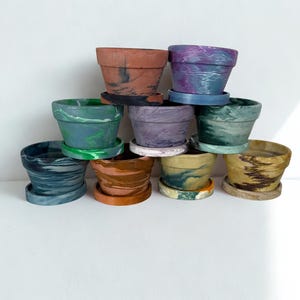 Może przedstawiać: Kolekcja małych, dekoracyjnych doniczek w różnych kolorach, w tym brązowym, fioletowym, zielonym i niebieskim. Każda doniczka ma pasujący spodek. Doniczki są ułożone w kształt piramidy na białym tle.