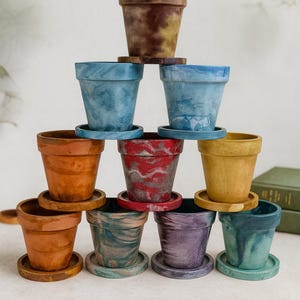 Può includere: Una collezione di piccoli vasi decorativi in vari colori, tra cui marrone, blu, rosso e giallo. Ogni vaso poggia su un piattino abbinato. I vasi sono impilati a forma di piramide, mostrando i loro diversi design.