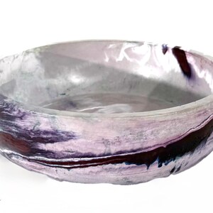May include: A round, shallow bowl featuring a marbled finish with swirls of light lavender, white, and deep dark purple. The surface has a smooth, glossy texture with a slightly uneven, organic rim and a wide, open interior basin design.