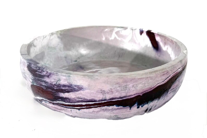 May include: A round, shallow bowl featuring a marbled finish with swirls of light lavender, white, and deep dark purple. The surface has a smooth, glossy texture with a slightly uneven, organic rim and a wide, open interior basin design.