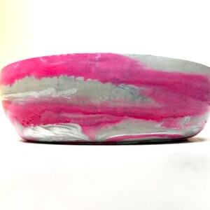 May include: A round, shallow bowl featuring a marbled pattern of vibrant hot pink and light gray. The exterior surface displays a smooth, swirling blend of these two colors, creating a continuous, fluid design around the entire circumference.