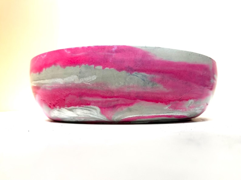 May include: A round, shallow bowl featuring a marbled pattern of vibrant hot pink and light gray. The exterior surface displays a smooth, swirling blend of these two colors, creating a continuous, fluid design around the entire circumference.