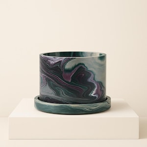 Könnte beinhalten: Ein zylindrischer Blumentopf mit passendem Untersetzer, der ein marmoriertes Muster in Grün-, Lila- und Grautönen aufweist. Der Topf steht auf einer weißen Oberfläche und unterstreicht sein einzigartiges Design. Ideal für Zimmerpflanzen.