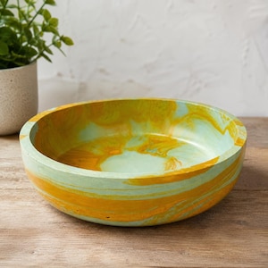 May include: A round, shallow bowl featuring a marbled pattern of vibrant yellow and soft mint green colors. The bowl has a smooth, solid construction with a wide, flat base and slightly curved sides, showcasing a swirling blend of these two colors.