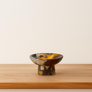 May include: A small, decorative ceramic bowl with a pedestal base. The bowl features a marbled design with shades of brown, yellow, and blue. The bowl is sitting on a wooden surface.
