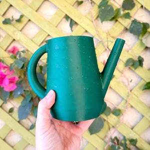 Op de afbeelding: Een groene gieter met een gebogen handvat en een lange tuit. De gieter wordt in een hand gehouden, met op de achtergrond een groen trellis en roze bloemen.