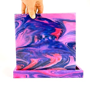 Puede incluir: Un panel cuadrado plano y una bandeja rectangular con un patrón jaspeado de tonos arremolinados en rosa vibrante, morado intenso y azul oscuro. Las superficies muestran un acabado liso y brillante con transiciones de color fluidas.