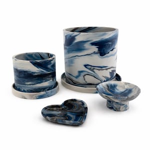 May include: A selection of ceramic homeware items featuring a marbled blue, white, and grey pattern. The set includes two cylindrical planters with saucers, a small heart-shaped dish, and a pedestal bowl. The pieces have a contemporary, artistic design.