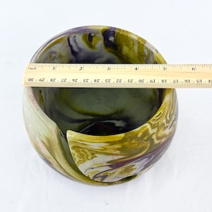 A ceramic yarn bowl look with the substantial weight and feel of cement - smooth and glossy finish. the ruler is set on top of the bowl to show a diameter of about 5 inches. the colors are marble and swirled in muted mustard yellow purple and white