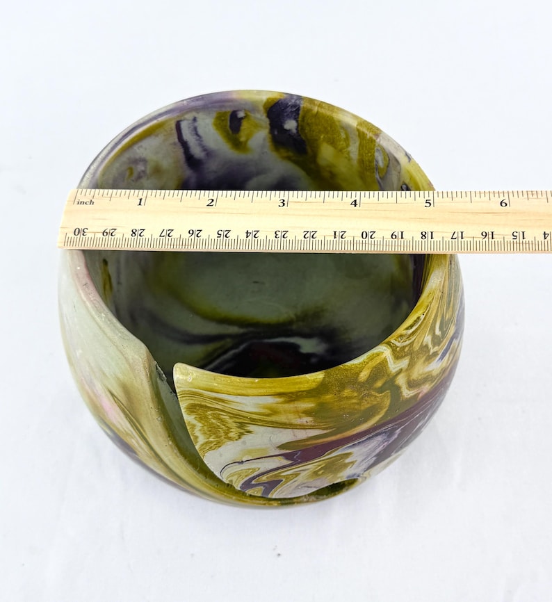 A ceramic yarn bowl look with the substantial weight and feel of cement - smooth and glossy finish. the ruler is set on top of the bowl to show a diameter of about 5 inches. the colors are marble and swirled in muted mustard yellow purple and white