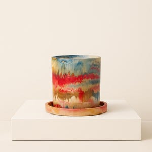 Può includere: Un vaso cilindrico in ceramica con un sottovaso abbinato. Il vaso presenta un design astratto colorato con striature rosse, blu e oro. Il vaso è su una superficie bianca.
